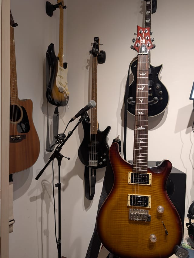 Mehrere Gitarren an einer Wand: eine Akustikgitarre, eine Stratocaster, ein Bass und eine PRS im Vordergrund