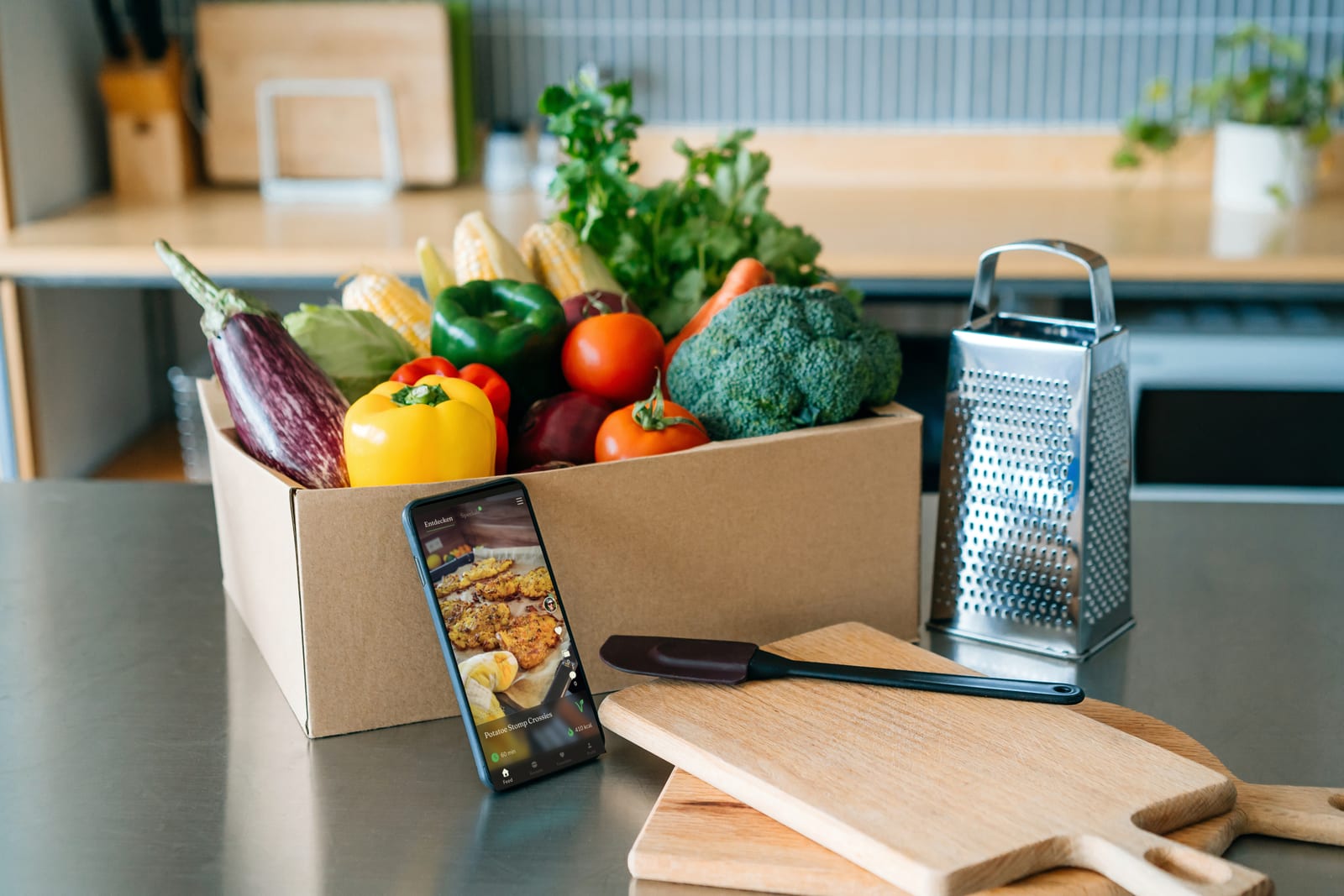 Ein Smartphone mit einem Rezept der Royal-Food-Club-App steht neben einer Kiste frischem Gemüse auf einer Küchenarbeitsfläche